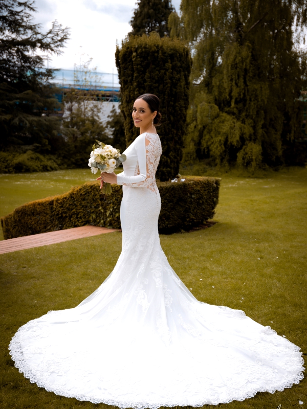 Full Length Shot Of A Bride In A Long Sleeved White Gown With A Dramatic Circular Train Standing On A Green Lawn.